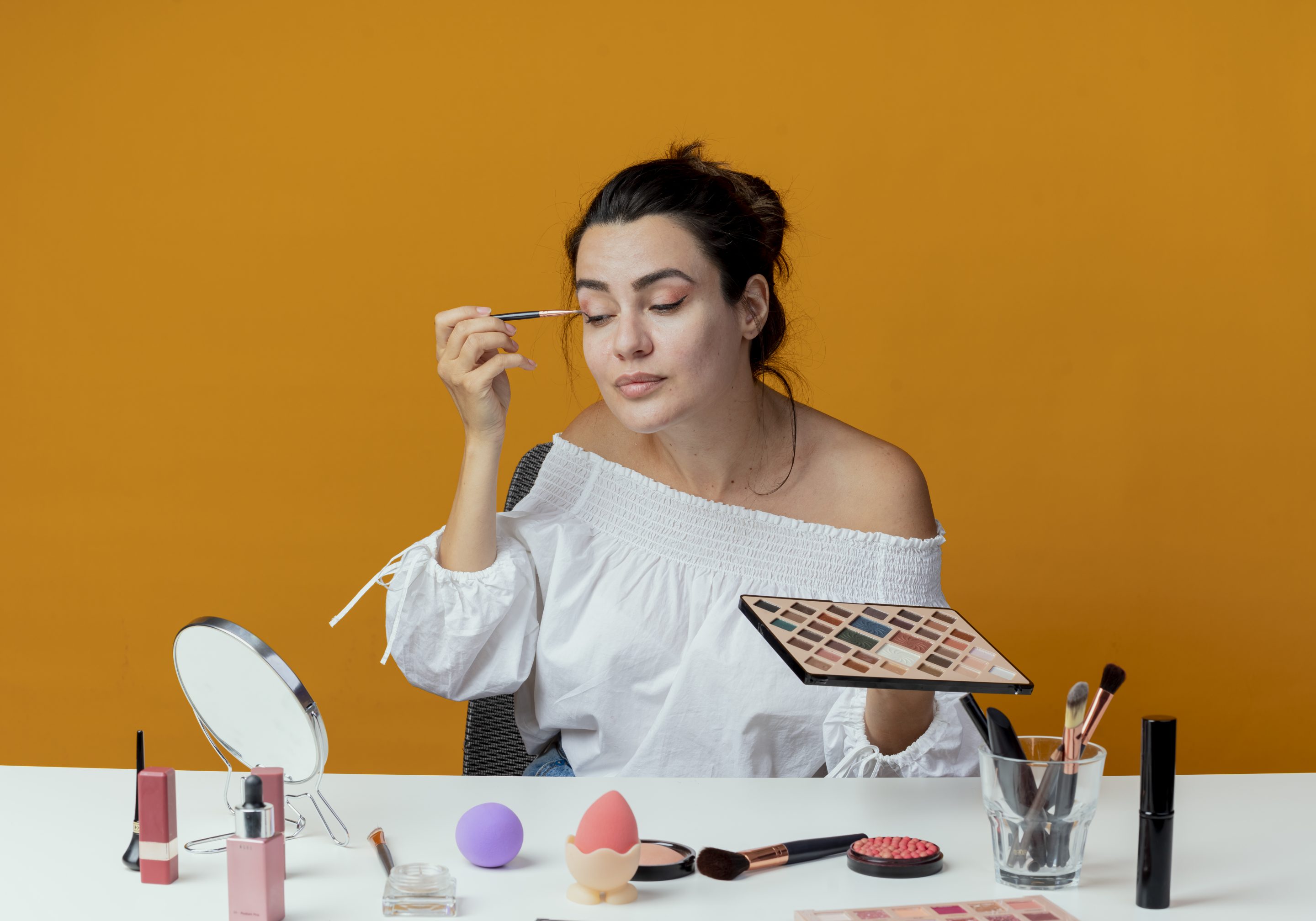 pleased beautiful girl sits at table with makeup tools looks at mirror applying eyeshadow with makeup brush isolated on orange background with copy space